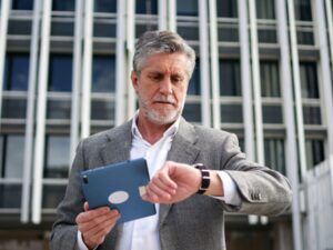 Gentlemen standing in front of an office tower, looking at his watch on one wrist and holding a tablet with the other.