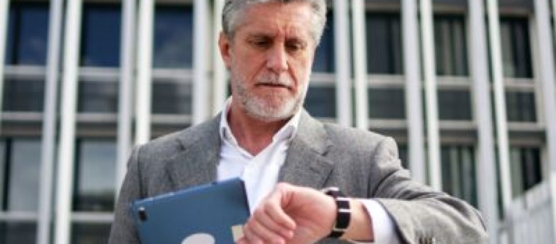 Gentlemen standing in front of an office tower, looking at his watch on one wrist and holding a tablet with the other.